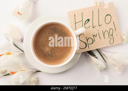 Carte avec mots HELLO SPRING, tasse de café et fleurs fraîches sur fond blanc, plat Banque D'Images