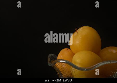 Gros plan d'un bol en verre contenant des prunes jaunes mûres fraîches avec un fond noir et un espace de copie Banque D'Images