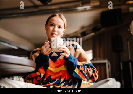 Jeune femme pensive portant un chandail coloré buvant du thé tout en étant assise à table dans le café Banque D'Images