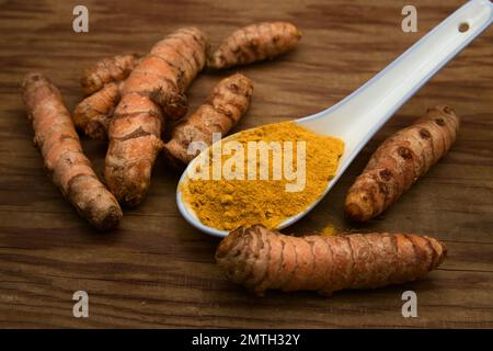 Une cuillère de poudre turmérique (Curcuma longa) entourée de rhizomes de la même plante sur une planche de bois Banque D'Images