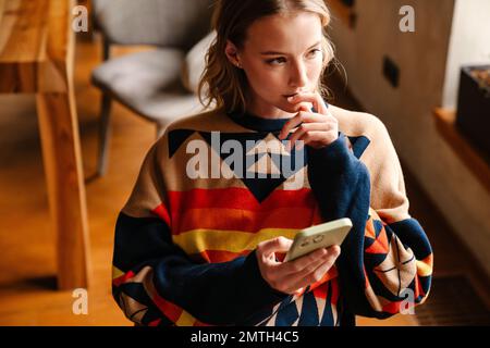 Jeune femme pensive portant un chandail coloré à l'aide d'un smartphone tout en étant assise dans un café Banque D'Images