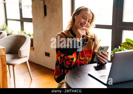 Jeune femme souriante dans des écouteurs ayant un appel vidéo via un smartphone tout en travaillant sur un ordinateur portable assis à une table dans un café Banque D'Images