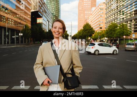 Jeune belle souriante heureuse femme d'affaires dans blazer avec ordinateur portable regardant l'appareil photo, tout en se tenant dans la rue dans la ville Banque D'Images