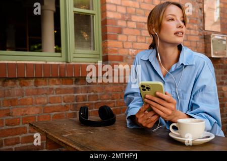 Jeune belle femme réfléchie avec la bouche ouverte dans les écouteurs tenant le téléphone et regardant de côté, tout en étant assis avec une tasse de café près de la brique de table Banque D'Images
