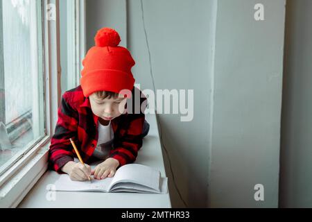 garçon fait ses devoirs. Le garçon se trouve sur le rebord de la fenêtre et écrit dans un bloc-notes. Un écolier fait ses devoirs par la fenêtre. Enseignement à domicile. Banque D'Images