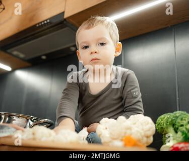 Un petit enfant aide à préparer le dîner depuis la vue du dessous. Bébé fils assis sur une table parmi des légumes frais mûrs. Une alimentation saine Banque D'Images