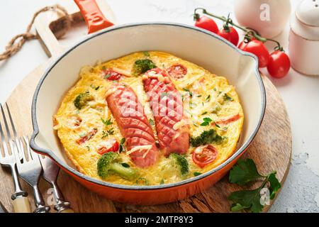 Omelette ou frittata avec deux saucisses, fromage fondu, tomates cerises, brocoli et persil sur poêle rouge ou poêle en fer sur fond blanc en béton Banque D'Images