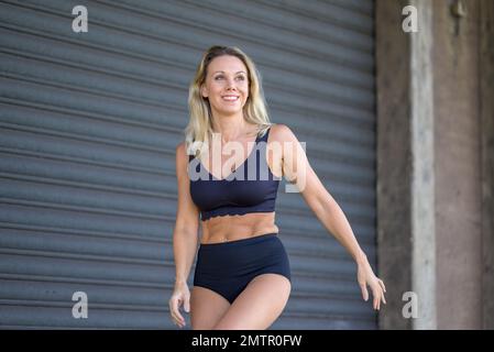 Belle femme blonde d'âge moyen portant un soutien-gorge noir et un pantalon noir souriant devant un volet gris Banque D'Images