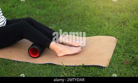 Journée mondiale de la santé. Une femme méconnaissable se réchauffe et travaille sur la structure des muscles du mollet avec un rouleau en mousse de massage sportif à l'extérieur sur l'herbe verte. Banque D'Images