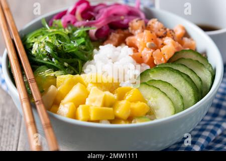 Un bol de poke avec du riz, du saumon, du concombre, de la mangue, de l'oignon, une salade de wakame, des graines de pavot et des graines de tournesol sur fond de bois Banque D'Images