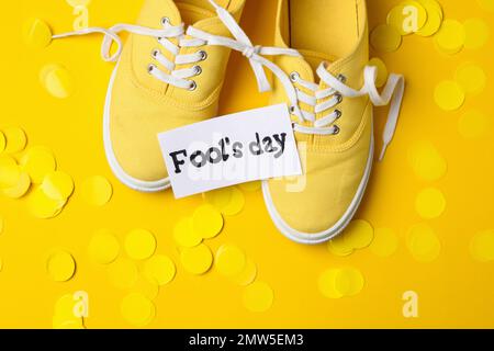 Chaussures attachées ensemble, note avec l'expression DE LA JOURNÉE DE L'IMBÉCILE et confetti sur fond jaune, couché à plat Banque D'Images