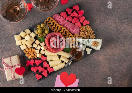 Carte de charcuterie pour la Saint-Valentin avec différentes variétés de saucisses, fromage, noix, olives et deux verres de vin rose sur fond marron, vue du dessus Banque D'Images