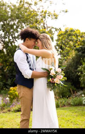 Vertical de la mariée et marié heureux et divers avec bouquet embrassant au mariage dans le jardin Banque D'Images