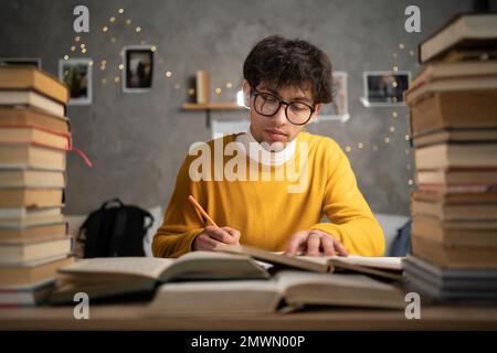 Concept d'éducation. Étudiant arabe étudiant en dortoir à une table avec des piles de livres, jeune homme se préparant pour les examens le soir en prenant des notes Banque D'Images