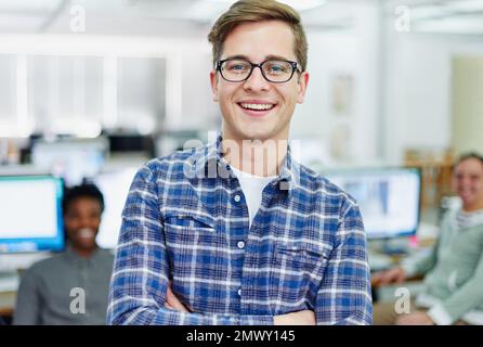 Aller dans l'entreprise. Portrait d'un jeune designer souriant debout dans un bureau avec des collègues travaillant en arrière-plan. Banque D'Images