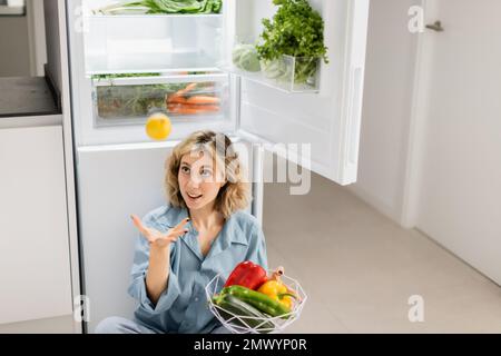 jeune femme assise près du réfrigérateur ouvert avec des légumes frais tout en jetant le citron dans l'air, image de stock Banque D'Images