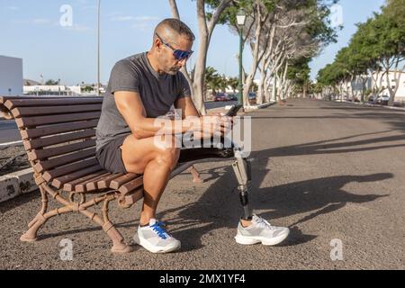 Toute la longueur de l'amputé mâle barbu avec prothèse de jambe assise sur le banc après avoir courez tout en parcourant le téléphone portable par beau temps Banque D'Images