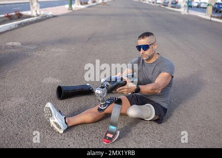 Par-dessus le jeune sportif déterminé en vêtements d'activité et lunettes de soleil assis sur la route tout en mettant sur la prothèse de jambe de lame avant l'entraînement dans la rue Banque D'Images