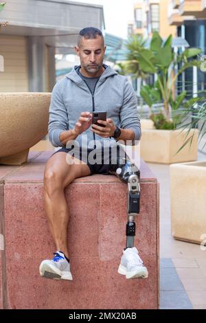 Un homme plein corps s'est amusé dans des vêtements de sport assis sur un bloc de marbre et parcourant les médias sociaux sur un téléphone mobile pendant une pause dans un entraînement de fitness en journée Banque D'Images