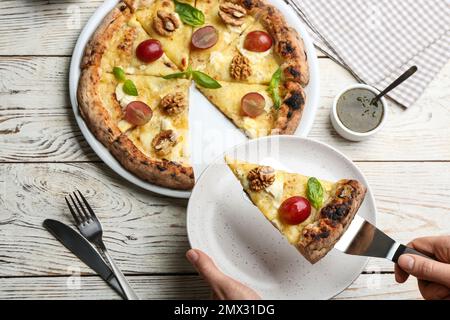 Femme prenant une tranche de pizza au fromage délicieux avec des noix et des raisins à une table en bois blanc, vue de dessus Banque D'Images