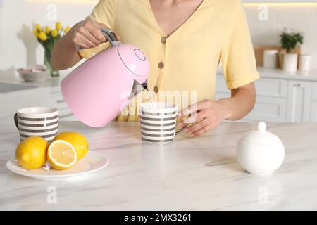 Femme versant de l'eau chaude de la bouilloire électrique moderne dans une tasse à l'intérieur, gros plan Banque D'Images