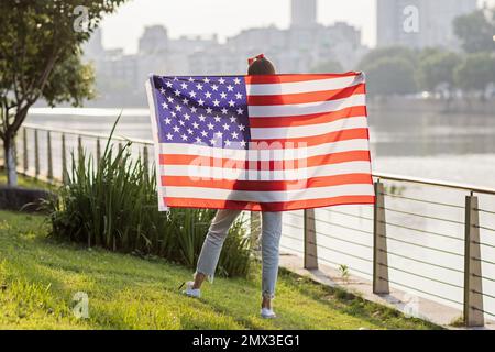 Vacances patriotiques. Bonne jeune femme caucasienne élégante avec drapeau américain à l'extérieur au coucher du soleil. Les Etats-Unis célèbrent l'indépendance le 4th juillet. Banque D'Images