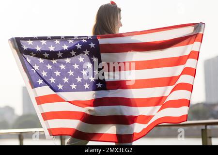 Vacances patriotiques. Bonne jeune femme caucasienne élégante avec drapeau américain à l'extérieur au coucher du soleil. Les Etats-Unis célèbrent l'indépendance le 4th juillet. Banque D'Images