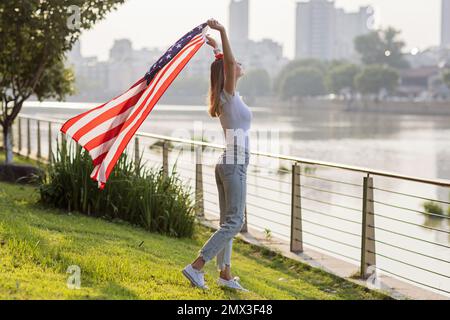 Vacances patriotiques. Bonne jeune femme caucasienne élégante avec drapeau américain à l'extérieur au coucher du soleil. Les Etats-Unis célèbrent l'indépendance le 4th juillet. Banque D'Images