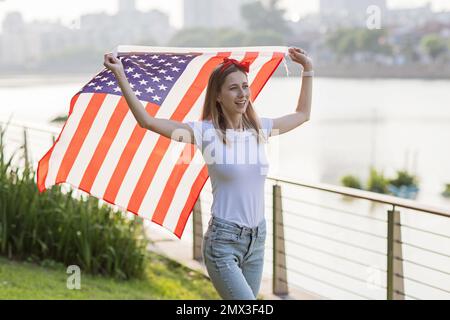 Vacances patriotiques. Bonne jeune femme caucasienne élégante avec drapeau américain à l'extérieur au coucher du soleil. Les Etats-Unis célèbrent l'indépendance le 4th juillet. Banque D'Images