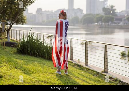 Vacances patriotiques. Bonne jeune femme caucasienne élégante avec drapeau américain à l'extérieur au coucher du soleil. Les Etats-Unis célèbrent l'indépendance le 4th juillet. Banque D'Images