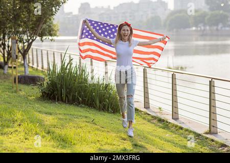 Vacances patriotiques. Bonne jeune femme caucasienne élégante avec drapeau américain à l'extérieur au coucher du soleil. Les Etats-Unis célèbrent l'indépendance le 4th juillet. Banque D'Images