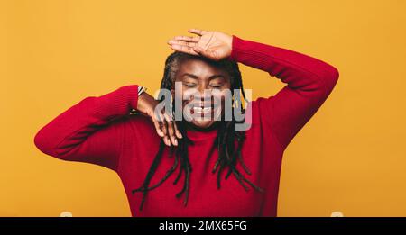 Bonne femme mûre riant et s'amusant tout en se tenant sur un fond jaune. Femme joyeuse avec des dreadlocks s'appréciant dans un studio. Banque D'Images
