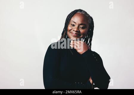 Bonne femme mûre avec des dreadlocks regardant la caméra avec un sourire dans un studio. Femme d'âge moyen à la mode debout sur fond gris. Banque D'Images