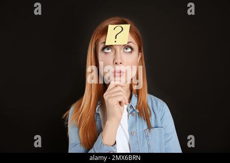 Femme pensive avec étiquette de point d'interrogation sur le front contre fond noir Banque D'Images