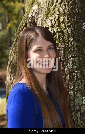 Un beau sourire illumine le visage d'une jeune femme. Elle a de longs cheveux et porte une robe bleue. Elle se tient à côté d'un arbre à l'extérieur. Banque D'Images