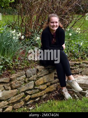 Un jeune adolescent est assis sur un mur de pierre dans un jardin. Elle se sourit et se embrasse à cause du froid du temps du début du printemps. Elle est habillée Banque D'Images