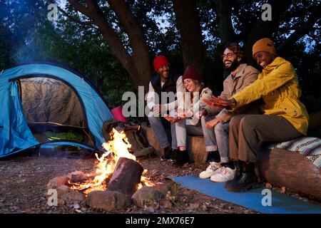 Un groupe multiethnique de jeunes amis campent à l'extérieur, assis autour d'un feu de camp sur la forêt. Banque D'Images