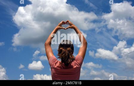 Jeune, femme, adolescent forme le coeur avec ses mains. Elle regarde dans un ciel bleu avec de grands nuages moelleux et exprime son amour et son appréciation f Banque D'Images