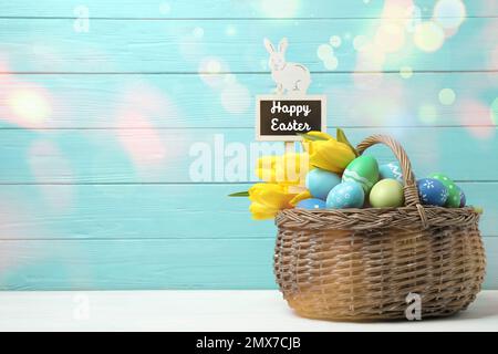 Œufs colorés, tulipes et étiquette avec texte Joyeux Pâques dans panier en osier sur table blanche sur fond bleu clair Banque D'Images