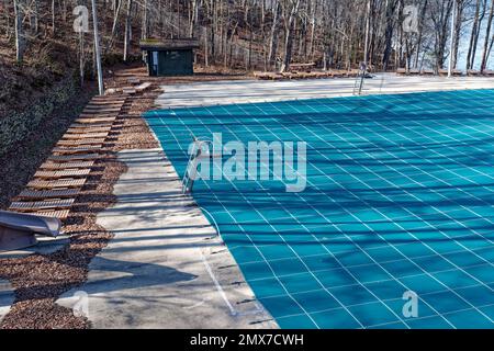 Arrière d'une grande piscine publique dans un parc fermé pour la saison avec une bâche couvrant l'eau et des meubles de salon sur la terrasse de ciment entouré par f Banque D'Images