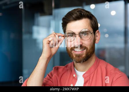 Portrait en gros plan de jeune homme d'affaires en chemise rouge, homme mature en lunettes souriant et regardant l'appareil photo dans le bureau près de la fenêtre, investisseur réussi heureux avec des réalisations et un bon investissement. Banque D'Images