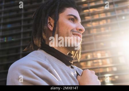 Vue latérale à angle bas d'un jeune homme joyeux avec des dreadlocks qui regardent loin tout en se tenant contre un bâtiment moderne Banque D'Images