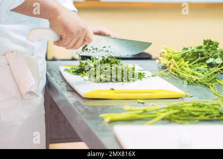 Crop anonyme femme chef en uniforme coupant le persil vert frais avec couteau sur la planche à découper dans la cuisine Banque D'Images