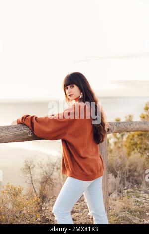 Vue latérale d'une femme voyageur avec de longs cheveux ondulés debout sur le point de vue tout en regardant l'appareil photo Banque D'Images