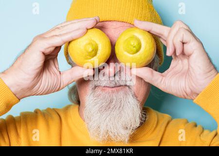 Homme âgé barbu en chandail jaune et chapeau couvrant les yeux avec des citrons sur fond bleu Banque D'Images