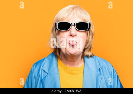 Portrait d'une femme âgée dans des lunettes de soleil tendance et des vêtements brillants regardant l'appareil photo tout en montrant la langue sur fond orange Banque D'Images