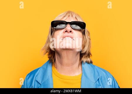 Gaie femme âgée en vêtements lumineux et lunettes de soleil souriant et regardant vers le haut tout en se tenant sur fond jaune Banque D'Images