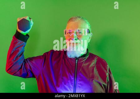 Hipster masculin senior barbu sérieux dans des vêtements décontractés et des lunettes debout sur fond vert tout en soulevant la haltère Banque D'Images