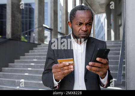 Un problème avec le compte. A contrarié un homme afro-américain dans un procès devant un bureau de banque. Il possède une carte de crédit. Il regarde avec véraison le téléphone, les cadrans, les chèques, les paiements. Banque D'Images