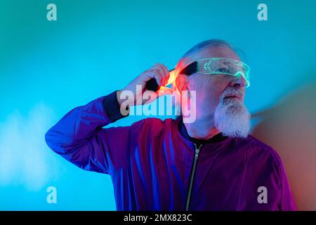 Culotte style hipster homme senior à barbe en lunettes et tenue décontractée avec vue sur le mur et lampe au néon en studio Banque D'Images